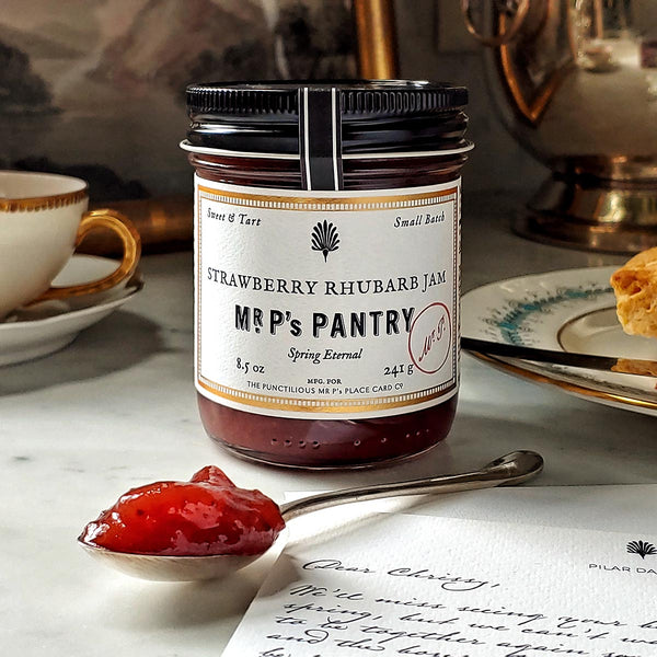 A jar of the Punctilious Mr. P's strawberry-rhubard jam on a marble tabletop with a porcelain tea cup and saucer, and biscuit in the background