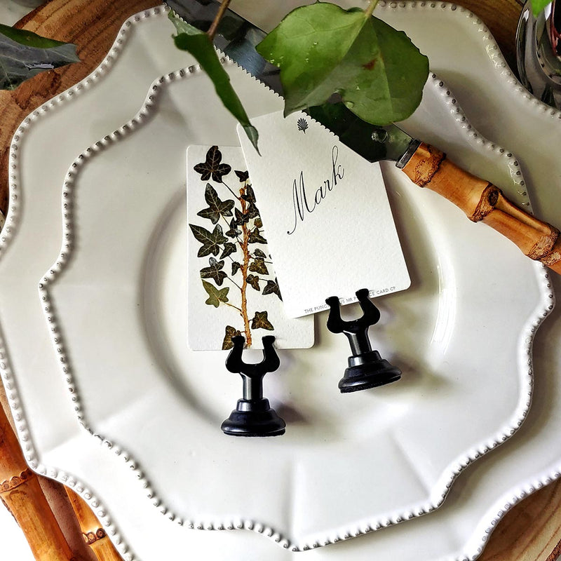 close up of The Punctilious Mr. P's Place Card Co. 'Ivy' custom place card set showing a name printed in digital calligraphy in spencerian font resting on white china with bamboo cutlery and live potted ivy in the background
