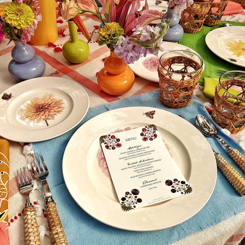 Marian McEvoy's mayfair size pressed poppy Custom  menu sits atop tablescape of Christopher Spitzmiller's dahlia dinner plates, wicker cutlery and red creel & gow tablecloth with beautiful bud vases and flowers