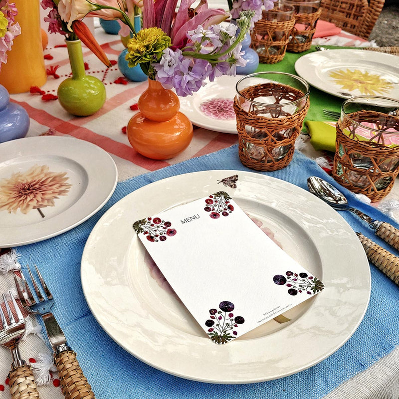 Marian McEvoy's mayfair size pressed poppy custom menu sits atop tablescape of Christopher Spitzmiller's dahlia dinner plates, wicker cutlery and red creel & gow tablecloth with beautiful bud vases and flowers